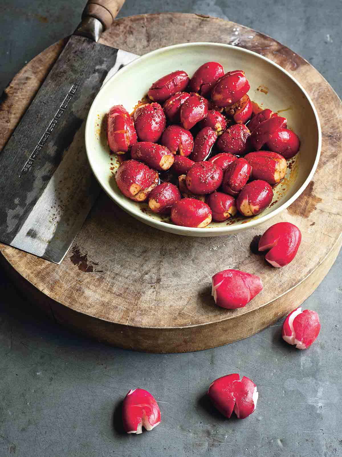 A white bowl of Radishes in chile oil sauce on a small round wooden table with a scraper beside the bowl.