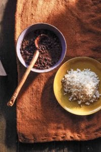 A bowl of Cuban black bean soup with a wooden ladle next to a yellow plate filled with white rice.