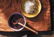 A bowl of Cuban black bean soup with a wooden ladle next to a yellow plate filled with white rice.