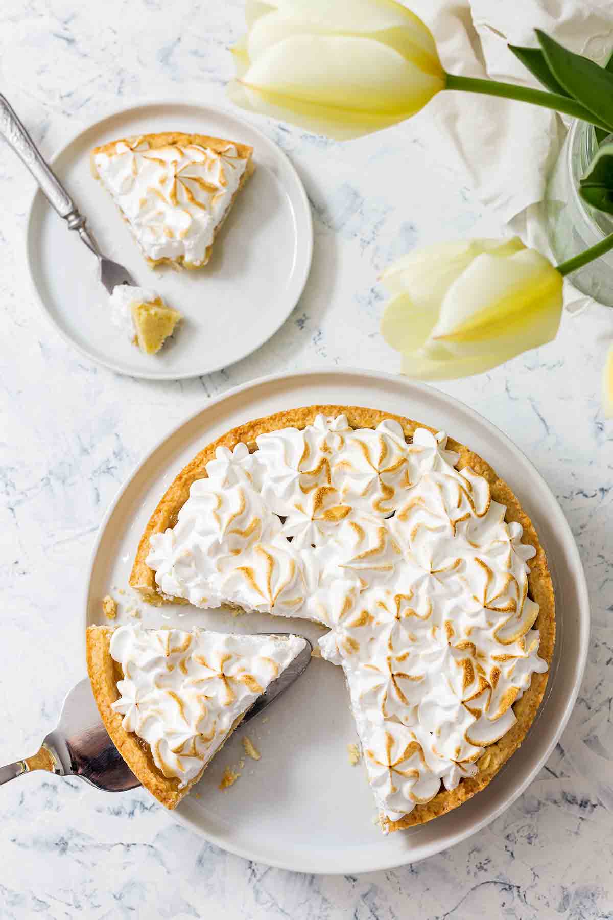 A lemon meringue tart in a white dish, with one slice on a plate beside it and one more being lifted out.