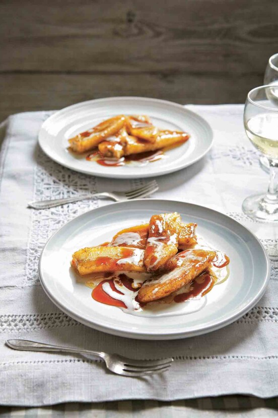 A white plate of pan-fried bananas with brandy that have been drizzle with coconut cream with a fork resting on either side.