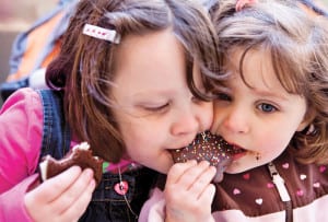 Two girls sharing a chocolate cookie decorated with sprinkles.