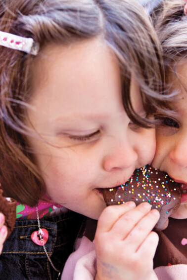 Two girls sharing a chocolate cookie decorated with sprinkles.