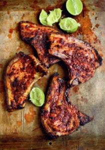 Four adobo marinated pork chops and several lime halves, some squeezed, on a wooden chopping block.