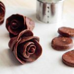 Dark chocolate taffy flowers sitting on a sheet of parchment, flanked with a cutter and a few chocolate petals.