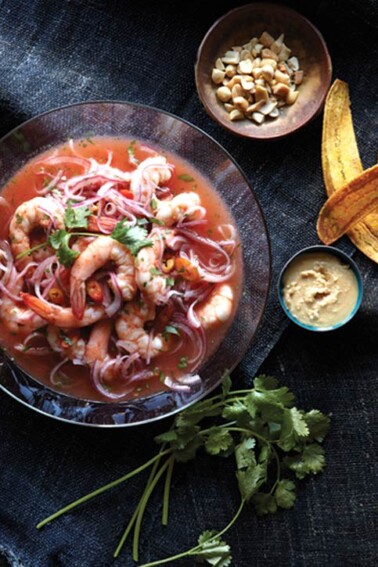 A big glass bowl filled with shrimp ceviche, flanked by a bowl of peanuts, a bunch of parsley, and a small bowl of onion relish.