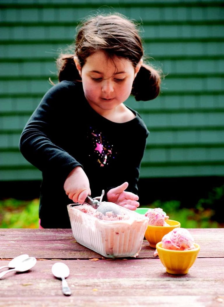 A little girl scooping strawberry ice cream into bowls.