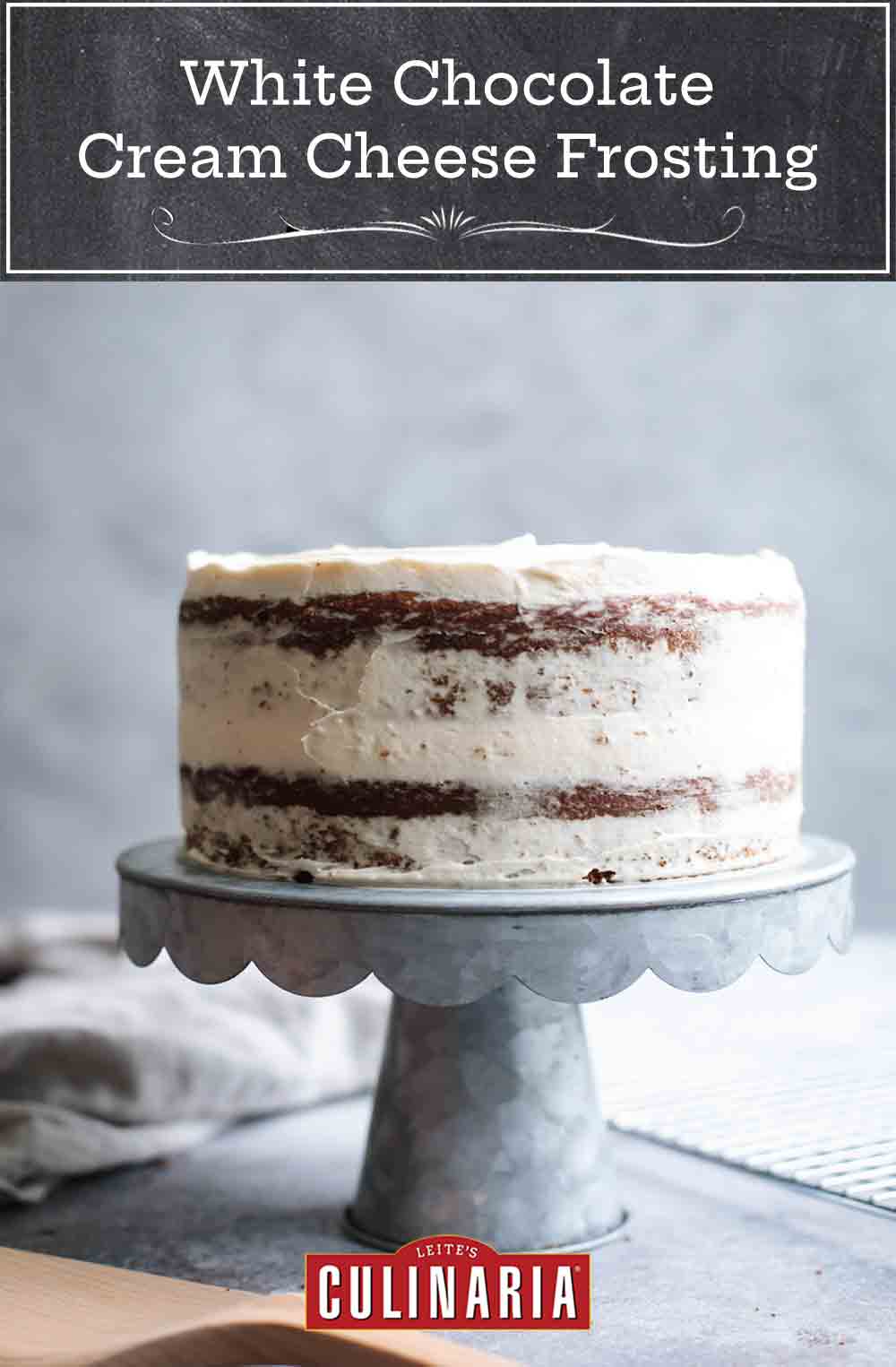 A double layer cake on a cake stand frosted in white chocolate-cream cheese frosting
