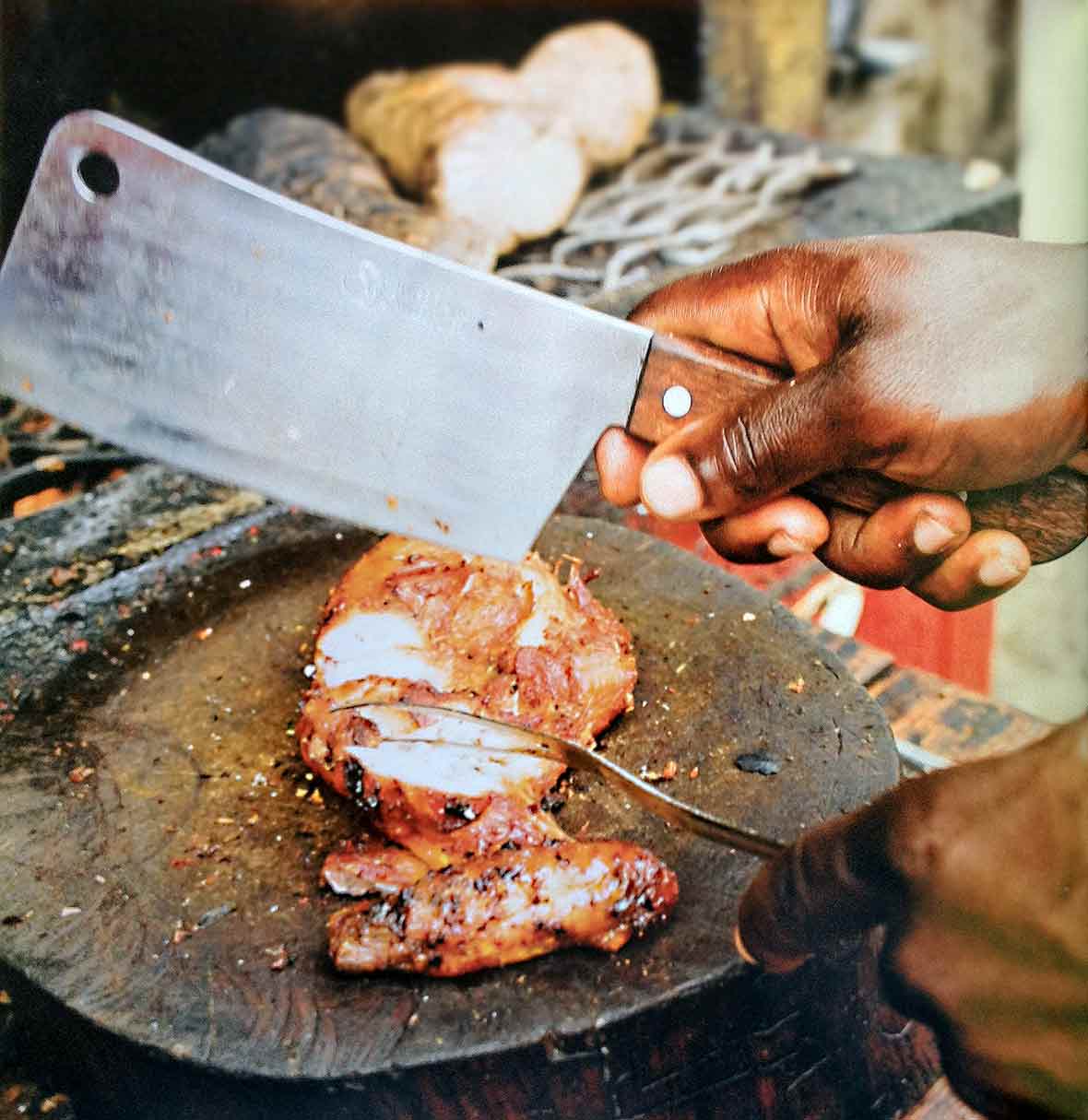 A sliced jerk chicken breast on a round cutting board, with a man's hand holding a cleaver above it