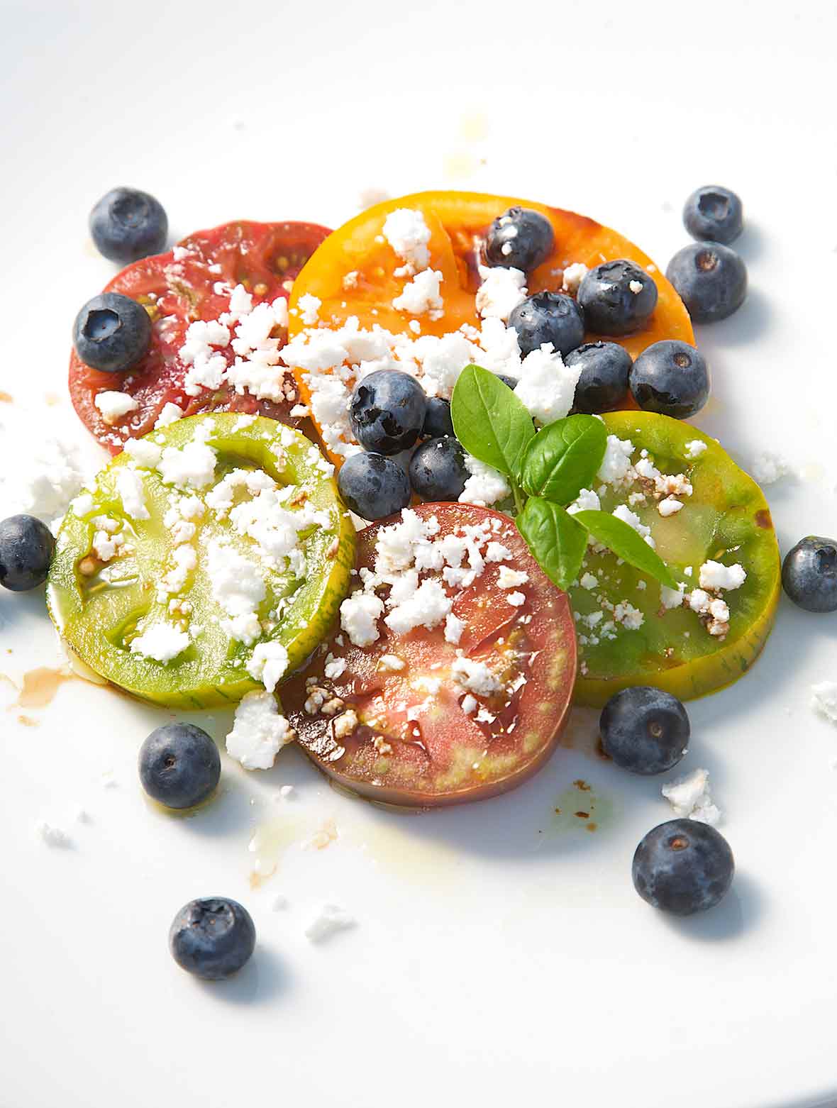 Sliced tomatoes with blueberries and crumbled feta scattered on top and a sprig of basil for garnish.