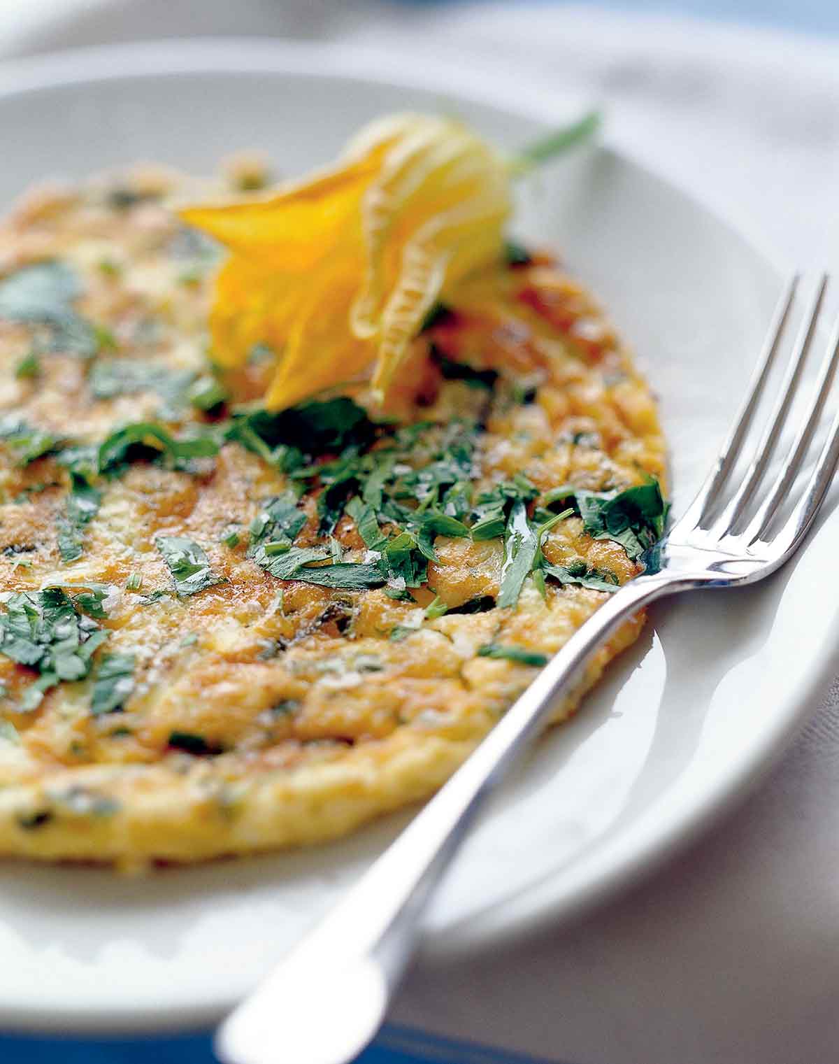An Italian frittata garnished with a squash blossom on a white plate with a fork resting beside it.