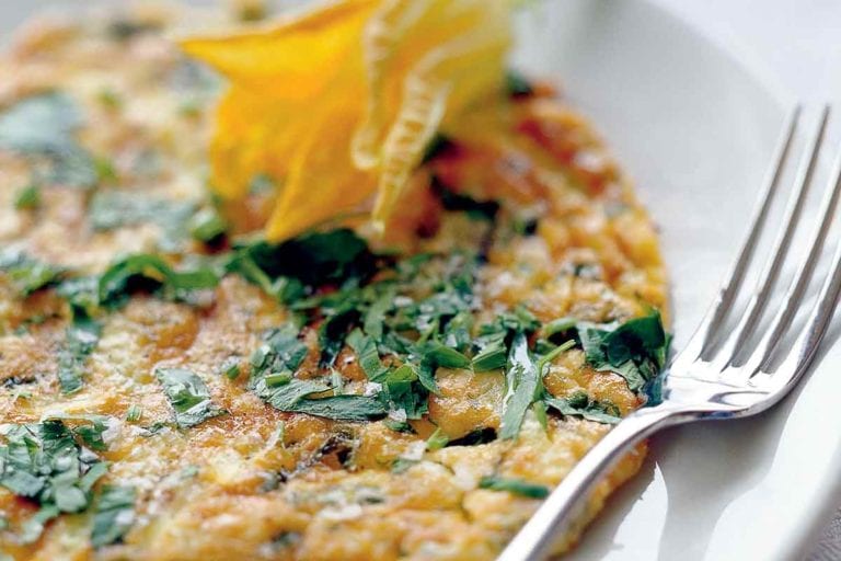 An Italian frittata garnished with a squash blossom on a white plate with a fork resting beside it.