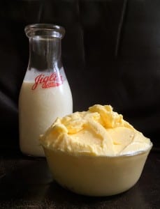 Homemade cultured butter mounded up in a yellow bowl with a glass jar of cream behind it.