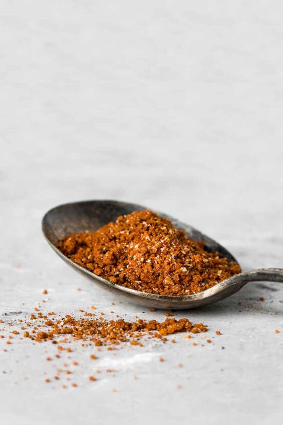 A spoon filled with Moroccan spice rub on a white surface with a little sprinkled beside the spoon.