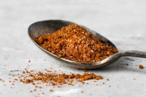 A spoon filled with Moroccan spice rub on a white surface with a little sprinkled beside the spoon.