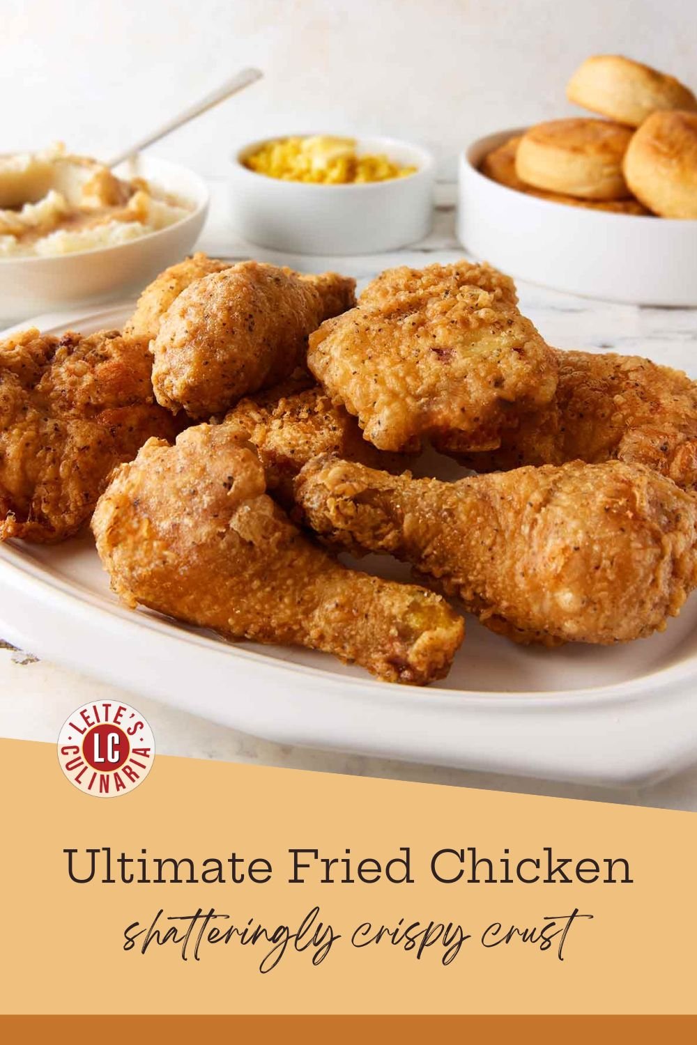 A white platter piled high with golden, crispy fried chicken, with classic side dishes of potatoes, corn, and biscuits behind it.