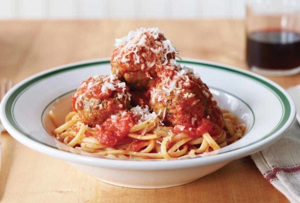 Three classic beef meatballs piled on pasta in a white and green bowl an finished with Parmesan.