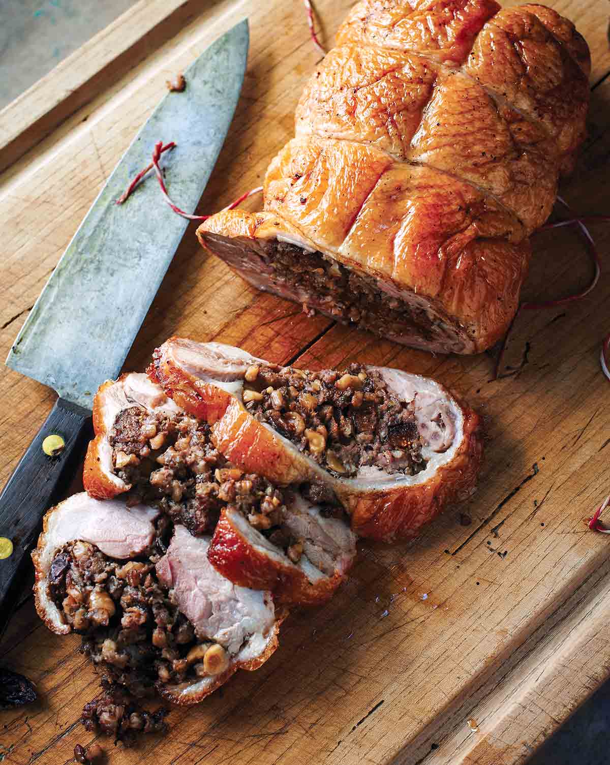 A roast duck stuffed with farro, figs, and hazelnuts, with three slices cut from it on a wooden cutting board with a knife resting beside it.