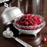 A silver and glass decorative dish filled with raw cranberry sauce. A lid for the dish and a silver spoon rest beside the dish.