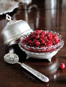 A silver and glass decorative dish filled with raw cranberry sauce. A lid for the dish and a silver spoon rest beside the dish.