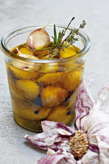 A pint-sized canning jar filled with garlic confit, sprigs of thyme and rosemary, and a partially separated head of garlic lying beside the jar.