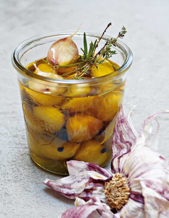 A pint-sized canning jar filled with garlic confit, sprigs of thyme and rosemary, and a partially separated head of garlic lying beside the jar.