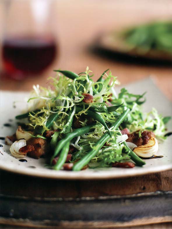 Haricots Verts with Frisée and Bacon on a white plate with a glass of red wine in the background.