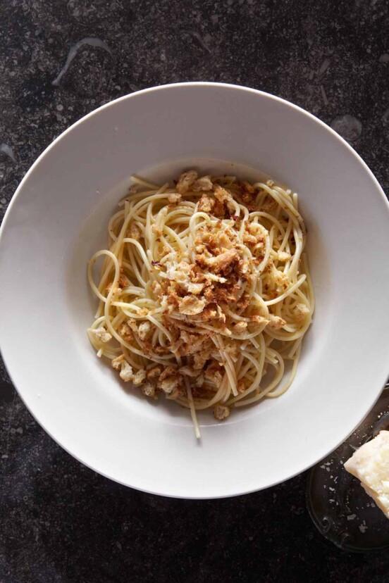 A white bowl filled with spaghetti topped with bread crumbs and a chunk of pecorino beside the bowl.