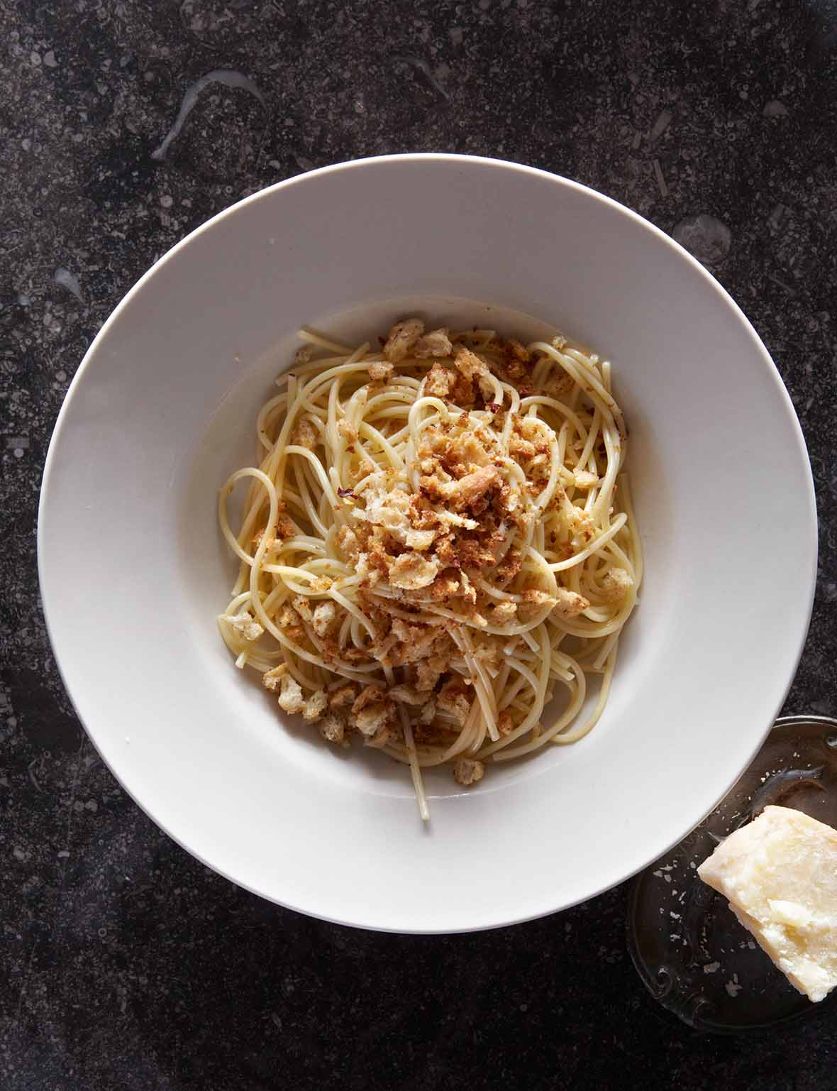 A white bowl filled with spaghetti topped with bread crumbs and a chunk of pecorino beside the bowl.