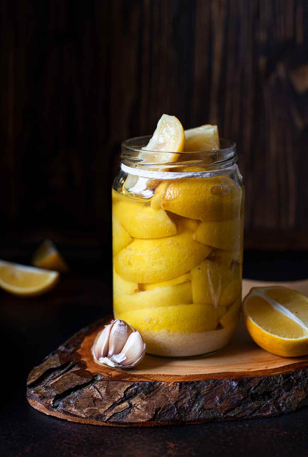 A jar of preserved lemons on a wooden board with lemon wedges and a few garlic cloves beside it.