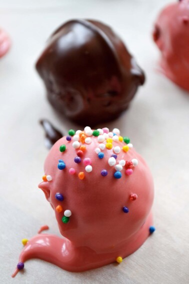 Cake pops covered with pink and chocolate icing and sprinkles, sitting on a white counter.