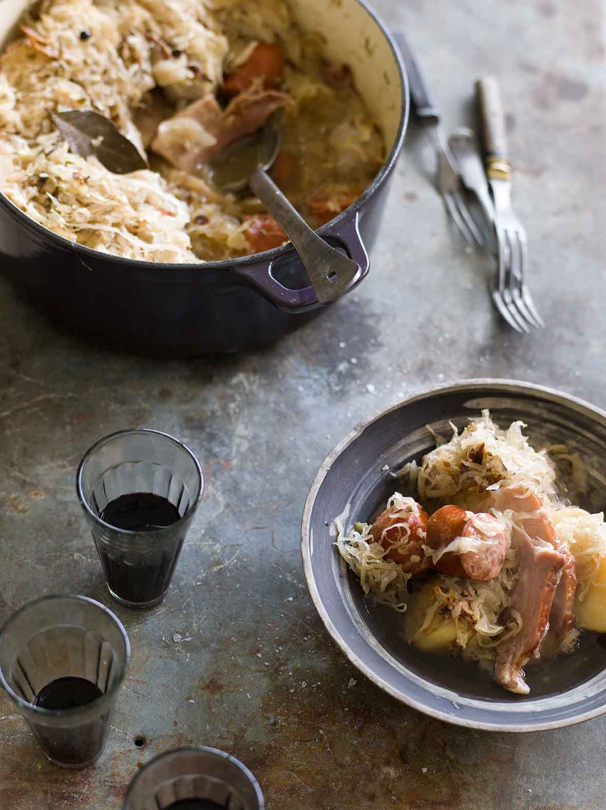 A Dutch oven and a porcelain bowl filled with choucroute garnie, made with pork, sausage, and sauerkraut, with two glasses of wine on the side.