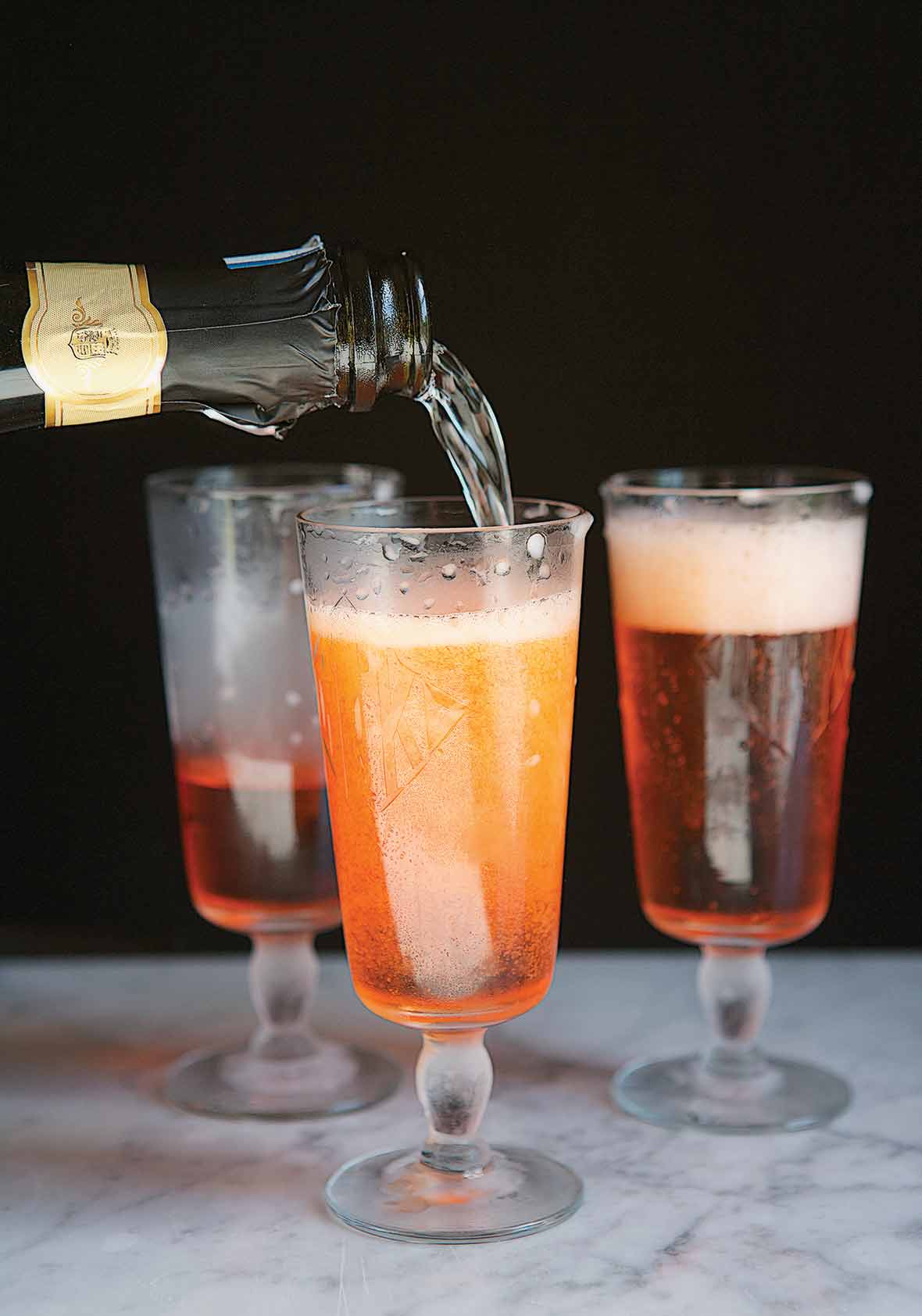 Prosecco being added to a Prosecco and Aperol cocktail, with two partially filled glasses behind it.