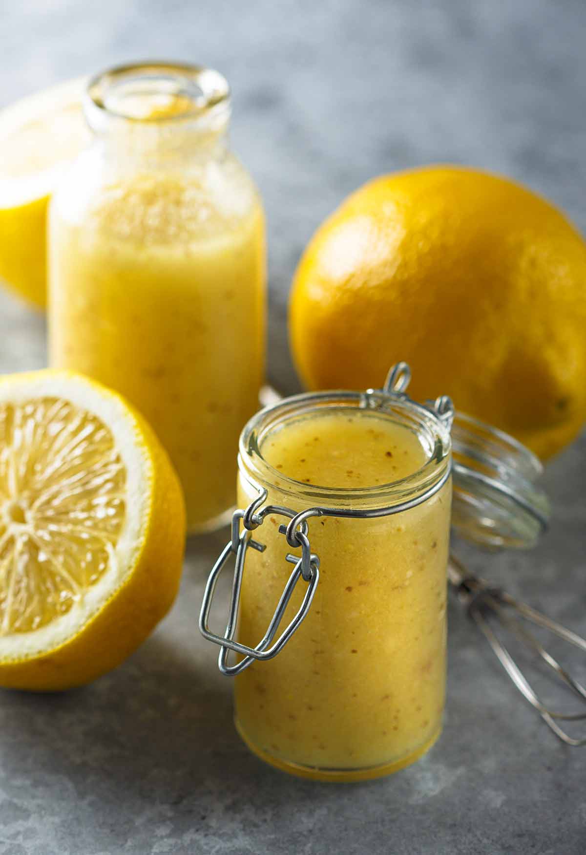 Two jars of lemon vinaigrette, two lemon halves, and a wire whisk.