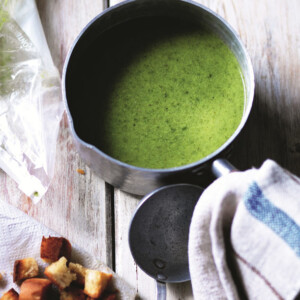 A saucepan filled with lettuce soup on a wooden table, with a dish cloth and a pile of toasted croutons beside it.