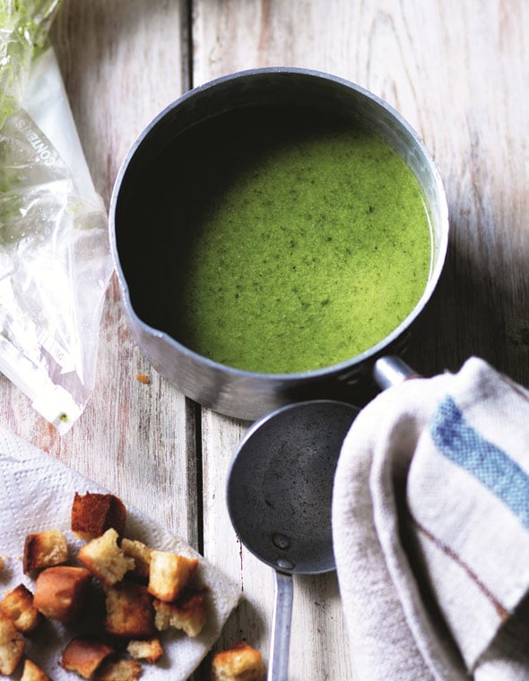 A saucepan filled with lettuce soup on a wooden table, with a dish cloth and a pile of toasted croutons beside it.