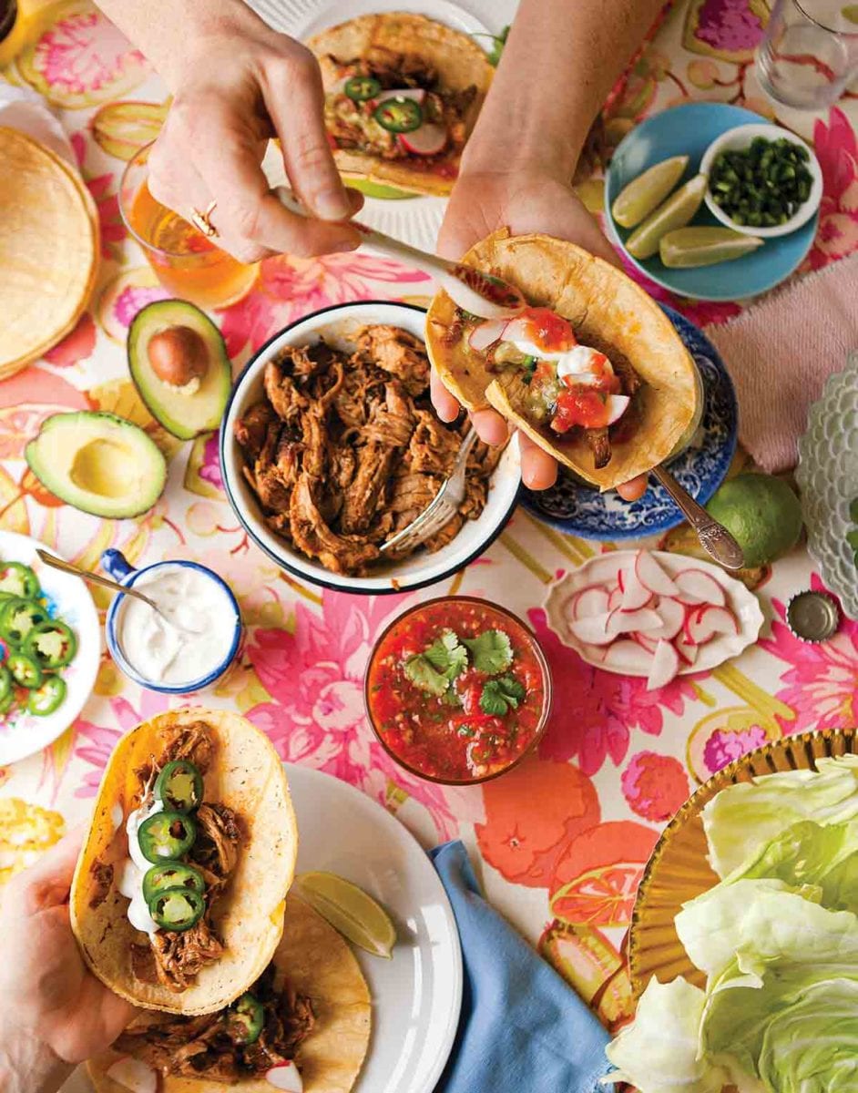 A person assembling a pork taco in a corn tortilla with a bowl of roast pork, a bowl of salsa, and a dish of sliced radishes on the table.