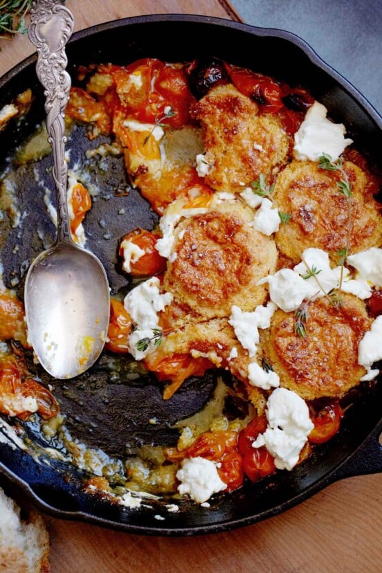 A cast-iron skillet half-filled with tomato and goat cheese cobbler and a spoon resting in the skillet.