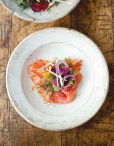 A white plate with salmon, fennel, and apple salad topped with red onion and dill.