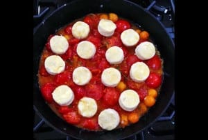 A cast-iron skillet filled with tomatoes being cooked for a tomato and goat cheese cobbler, topped with rounds of goat cheese.