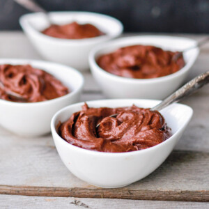 Four individual white bowls of avocado chocolate pudding with spoons on a wooden table.