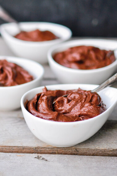 Four individual white bowls of avocado chocolate pudding with spoons on a wooden table.