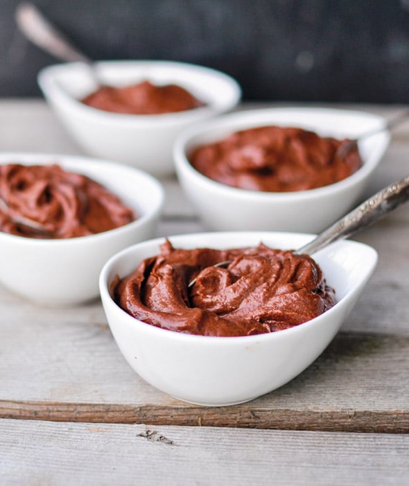 Four individual white bowls of avocado chocolate pudding with spoons on a wooden table.