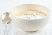 A white bowl of cauliflower soup on a white linen cloth with a spoon resting beside it.