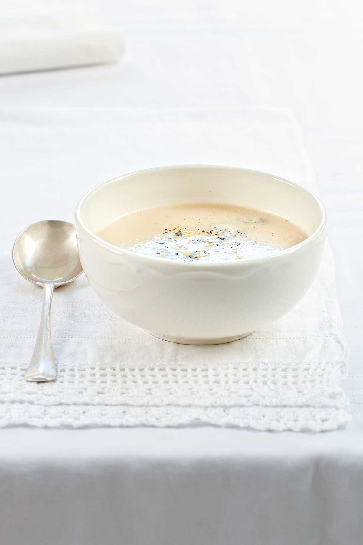 A white bowl of cauliflower soup on a white linen cloth with a spoon resting beside it.