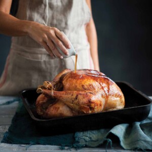 A woman pouring maple syrup over a maple glazed turkey in a black roasting pan.