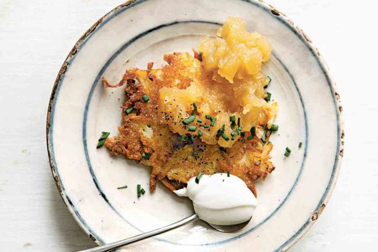 A cheddar and potato latke topped with chives and applesauce on a plate with a spoonful of sour cream beside the latke.