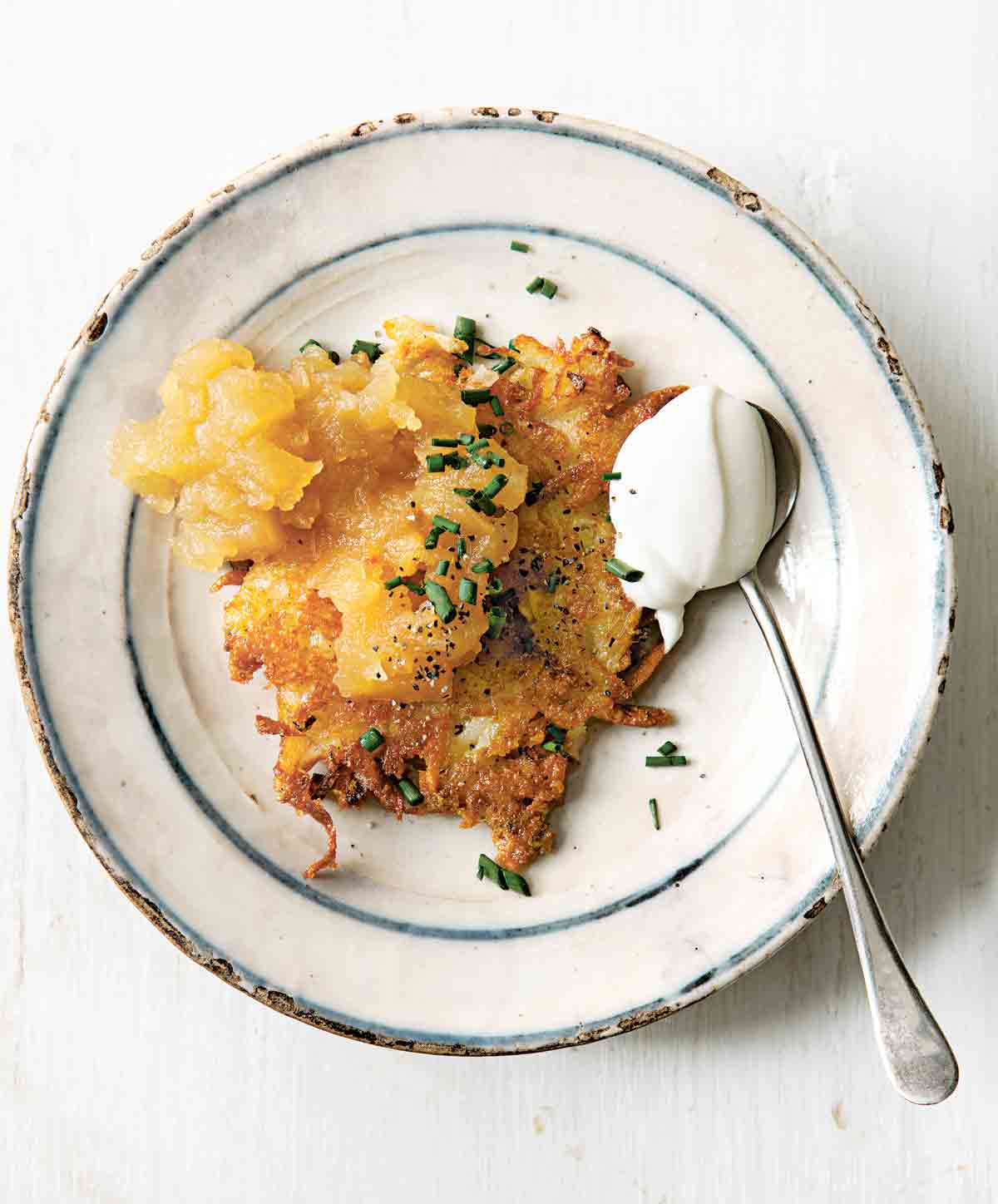 A cheddar and potato latke topped with chives and applesauce on a plate with a spoonful of sour cream beside the latke.