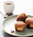Three German Berliners, or jelly doughnuts, with raspberry jam spilling out on a plate with a cup of coffee.