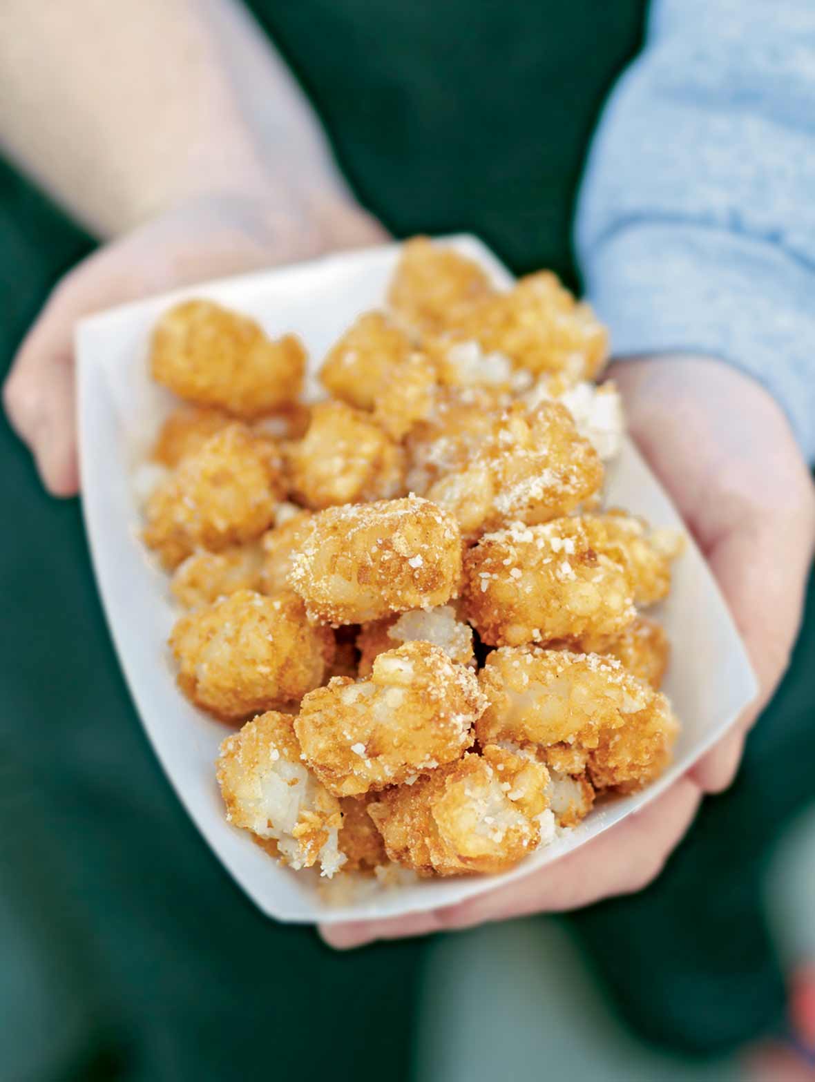 Two hands holding a paper container of tater tots sprinkled with Parmesan cheese